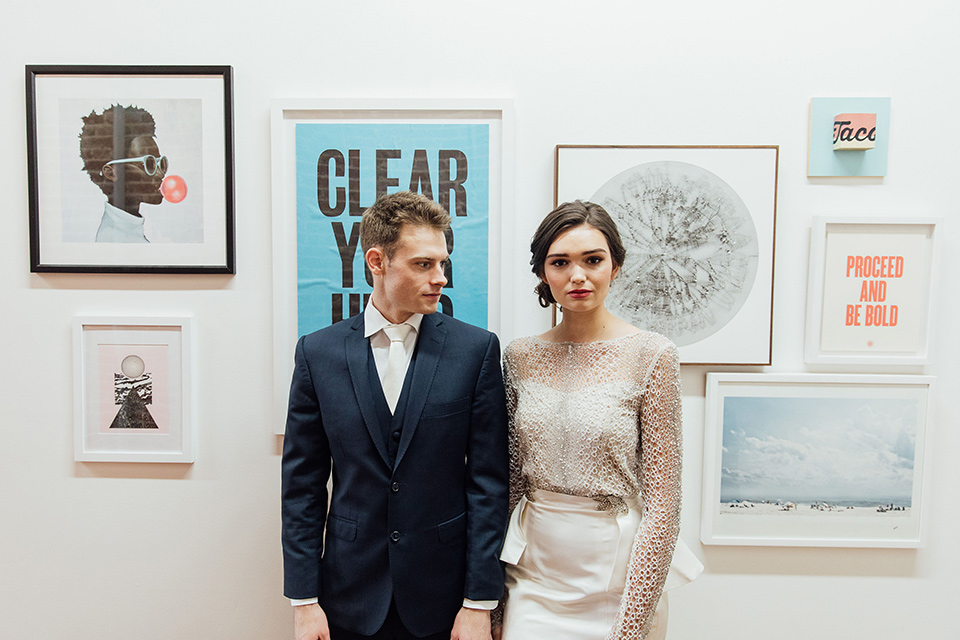  bride in a modern gown with a high neckline, no sleeves, and a tiered ruffled skirt and the groom in a light grey peak lapel suit with a coral long tie, in room gallery wall