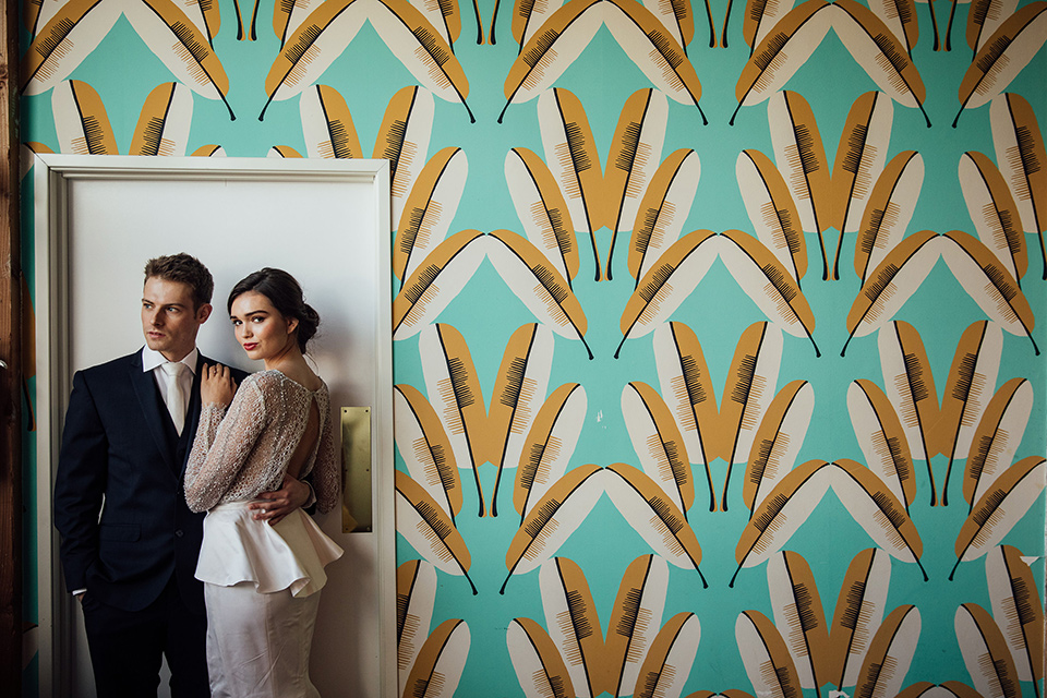  bride in a modern gown with a high neckline, no sleeves, and a tiered ruffled skirt and the groom in a light grey peak lapel suit with a coral long tie, in room gallery wall