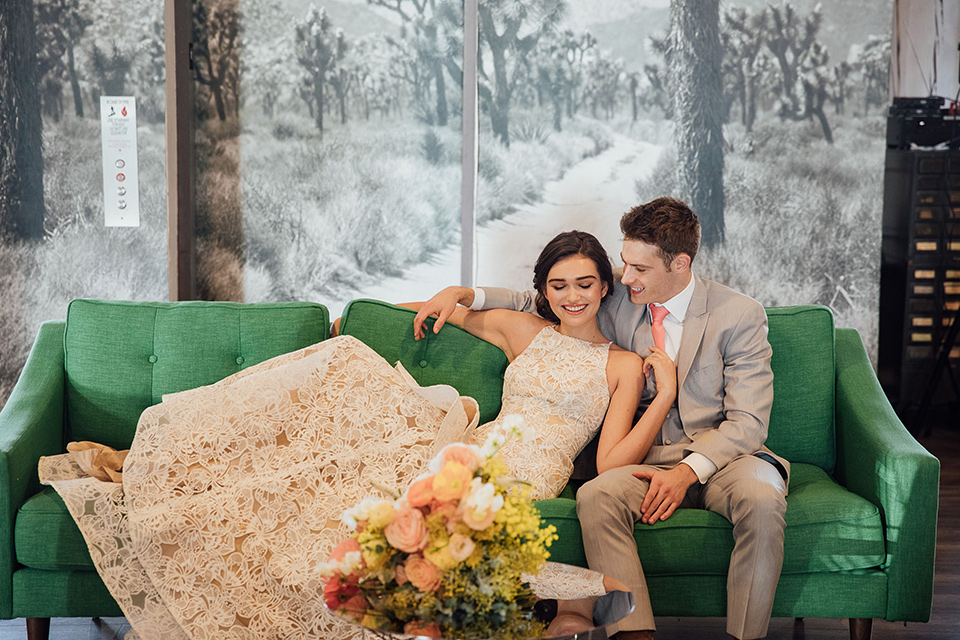  bride in a modern gown with a high neckline, no sleeves, and a tiered ruffled skirt and the groom in a light grey peak lapel suit with a coral long tie, in room gallery wall