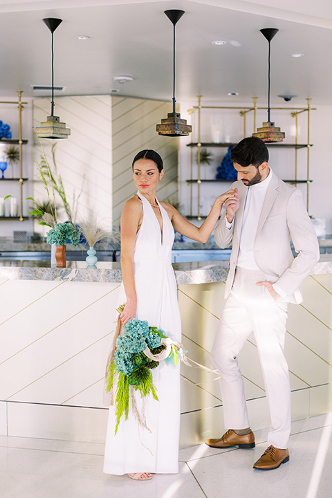  bride in a white jumpsuit with a halter and deep v neckline and her hair in a tight low bun and the groom in a light tan suit with a white shirt, next to the bar