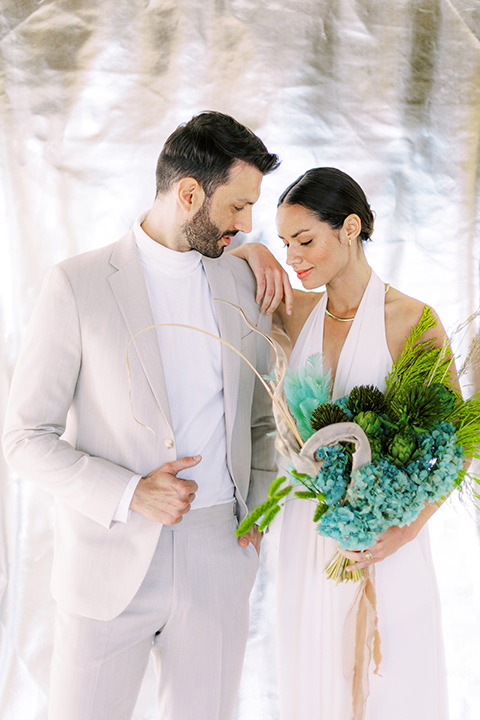  bride in a white jumpsuit with a halter and deep v neckline and her hair in a tight low bun and the groom in a light tan suit with a white shirt, at ceremony space