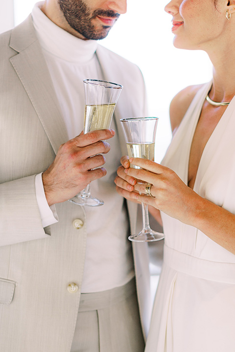  bride in a white jumpsuit with a halter and deep v neckline and her hair in a tight low bun and the groom in a light tan suit with a white shirt, cheersing with champagne