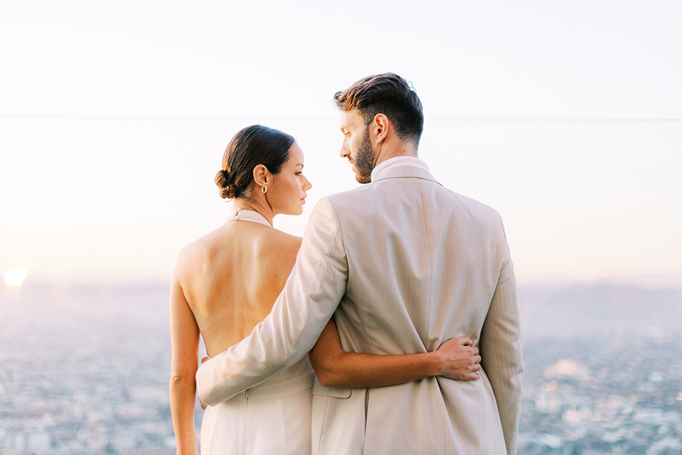  bride in a white jumpsuit with a halter and deep v neckline and her hair in a tight low bun with the city behind them