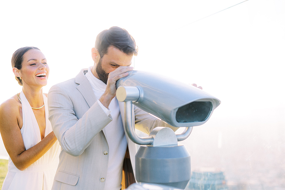  bride in a white jumpsuit with a halter and deep v neckline and her hair in a tight low bun with the city behind them looking through the telescope