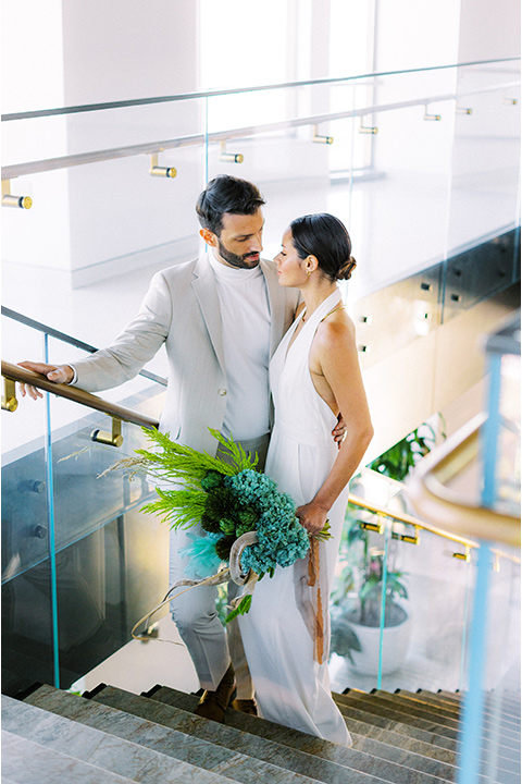  bride in a white jumpsuit with a halter and deep v neckline and her hair in a tight low bun and the groom in a light tan suit with a white shirt walking down the stairs