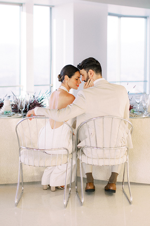  bride in a white jumpsuit with a halter and deep v neckline and her hair in a tight low bun and the groom in a light tan suit with a white shirt sitting at the reception