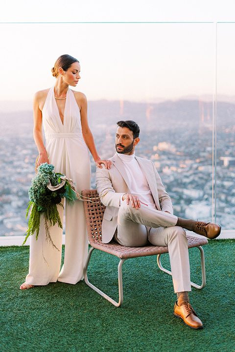  bride in a white jumpsuit with a halter and deep v neckline and her hair in a tight low bun and the groom in a light tan suit with a white shirt sitting out on the rooftop with the city behind them