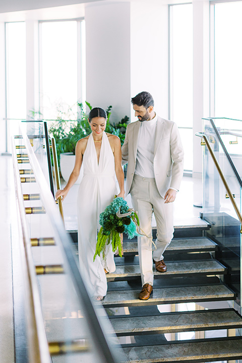  bride in a white jumpsuit with a halter and deep v neckline and her hair in a tight low bun and the groom in a light tan suit with a white shirt, heading down the stairs