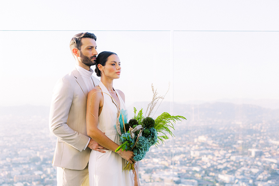  bride in a white jumpsuit with a halter and deep v neckline and her hair in a tight low bun with the city behind them