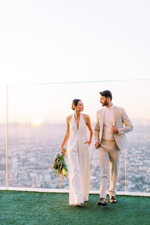  bride in a white jumpsuit with a halter and deep v neckline and her hair in a tight low bun and the groom in a light tan suit with a white shirt walking outside with the city behind them