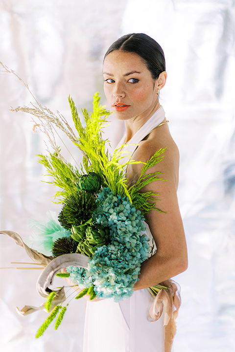  bride in a white jumpsuit with a halter and deep v neckline and her hair in a tight low bun, and carrying a blue and green floral bouquet