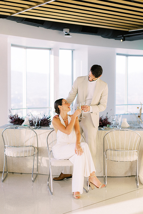  bride in a white jumpsuit with a halter and deep v neckline and her hair in a tight low bun and the groom in a light tan suit with a white shirt sitting at the reception table