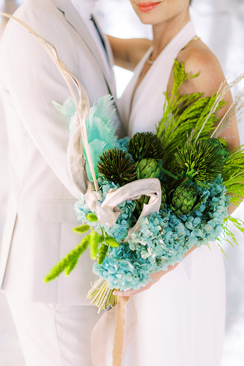  bride in a white jumpsuit with a halter and deep v neckline and her hair in a tight low bun and the groom in a light tan suit with a white shirt, close up on tropical style florals