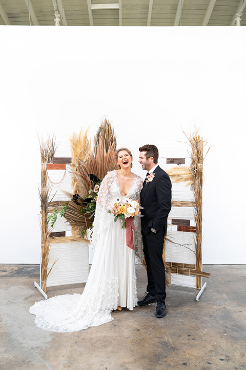  the bride in a lace white gown with long sleeves and a flowing skirt with hair in a modern low bun and the groom in a black suit with a long black tie next to the ceremony decor 