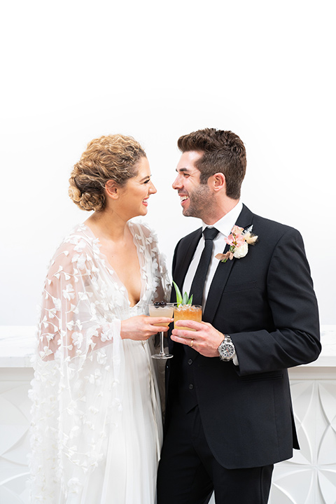  the bride in a lace white gown with long sleeves and a flowing skirt with hair in a modern low bun and the groom in a black suit with a long black tie with drinks 