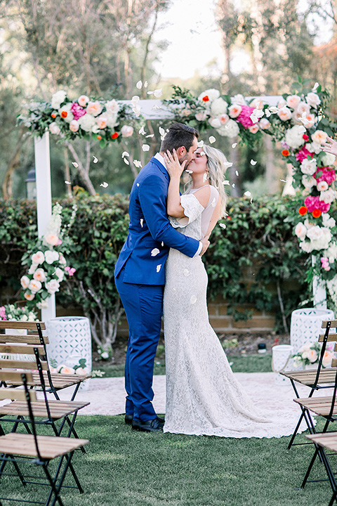  groom in a cobalt blue suit with a black long tie and brown shoes and the bride in a white bohemian gown with a high neckline and cold shoulder detail kissing