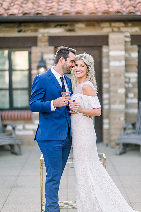  groom in a cobalt blue suit with a black long tie and brown shoes and the bride in a white bohemian gown with a high neckline and cold shoulder detail with champagne
