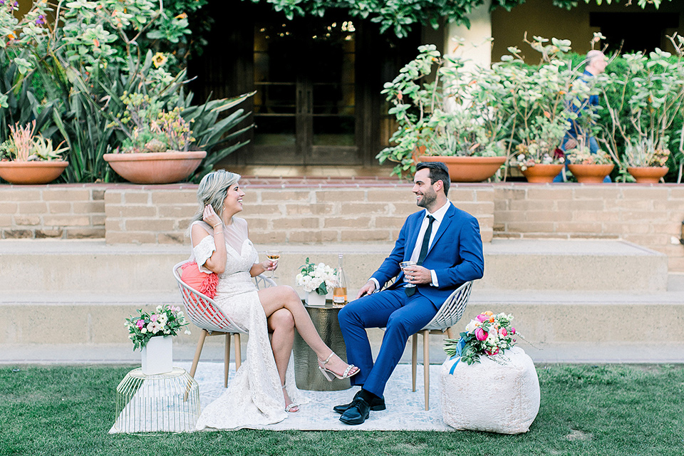  groom in a cobalt blue suit with a black long tie and brown shoes and the bride in a white bohemian gown with a high neckline and cold shoulder detail sitting at reception