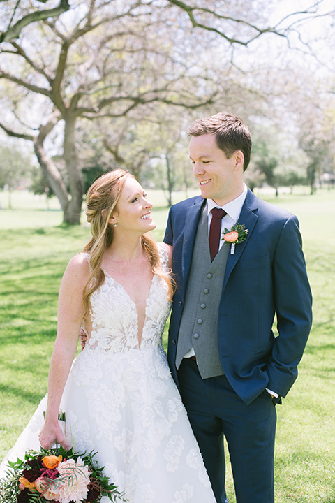  the bride in a flowing a line gown with a lace bodice in a low cut neckline and the groom in a dark blue suit with a grey vest and burgundy long tie