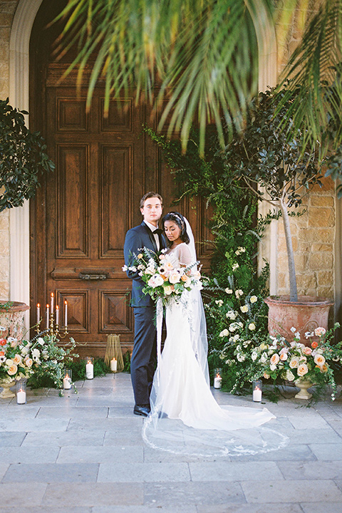  couple at ceremony groom in a navy blue tuxedo with a black bow tie and the bride in a white lace flowing gown and a long cathedral veil