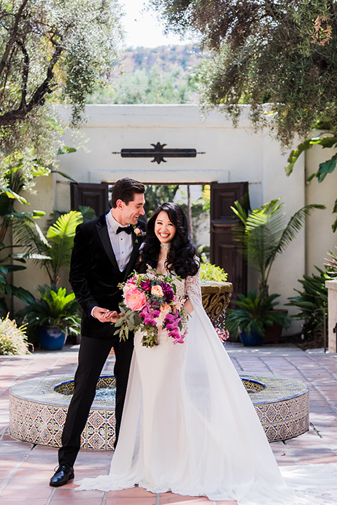  bride in a long gown with a flowing train and cape groom in a black velvet tuxedo with a black bow tie 