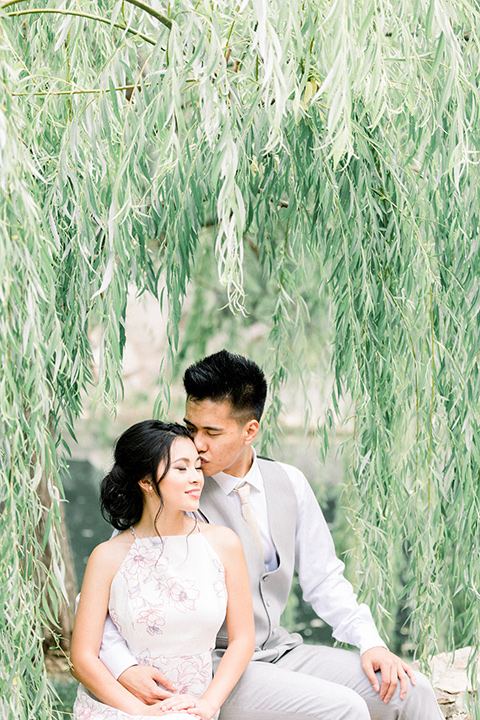 bride in a floral gown with a high neckline and a low bun hairstyle, the groom in a light grey pant and vest outfit with a white floral necktie together under the willow tree