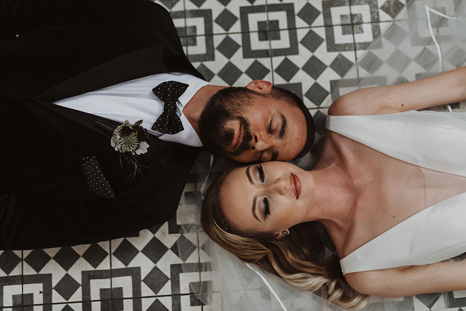  bride in a tulle a line gown with a sweetheart neckline and a crystal bodice and the groom in a black velvet tuxedo and black bow tie laying on ground