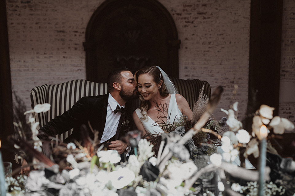  bride in a tulle a line gown with a sweetheart neckline and a crystal bodice and the groom in a black velvet tuxedo and black bow tie sitting at table