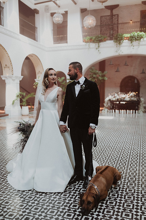  bride in a tulle a line gown with a sweetheart neckline and a crystal bodice and the groom in a black velvet tuxedo and black bow tie by reception table with dog 