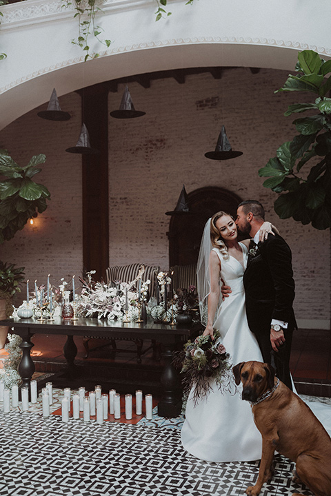  bride in a tulle a line gown with a sweetheart neckline and a crystal bodice and the groom in a black velvet tuxedo and black bow tie by reception table with dog 