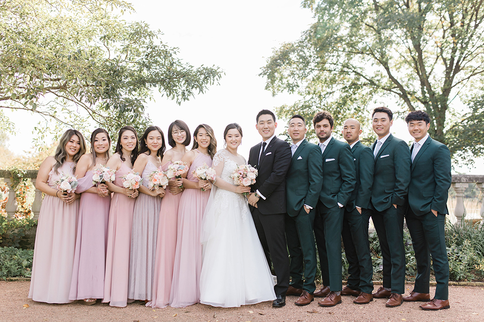  bride in a tulle ballgown with a plunging neckline and a loose bun, bridesmaids in light pink gowns, groomsmen in dark green suits and groom in a black suit with a black long tie