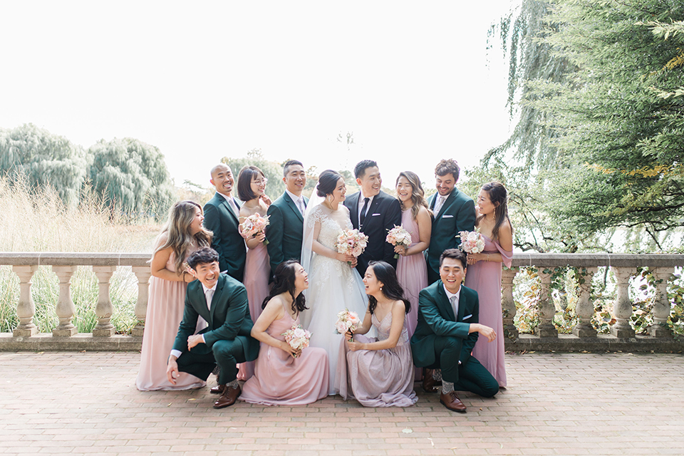  bride in a tulle ballgown with a plunging neckline and a loose bun, bridesmaids in light pink gowns, groomsmen in dark green suits and groom in a black suit with a black long tie