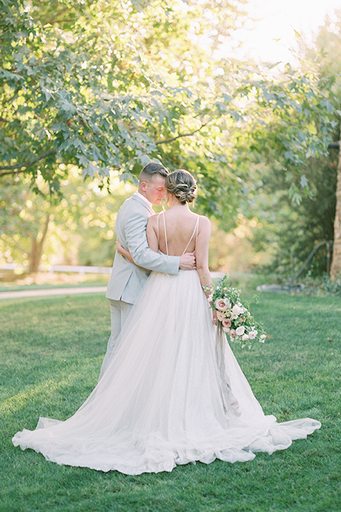  bride in a blush tulle ballgown with thin straps and hair up in a loose bun and the groom in a light grey suit with a white tie holding each other