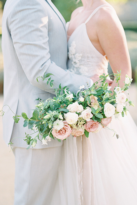  bride in a blush tulle ballgown with thin straps and hair up in a loose bun and the groom in a light grey suit with a white tie holding each other close