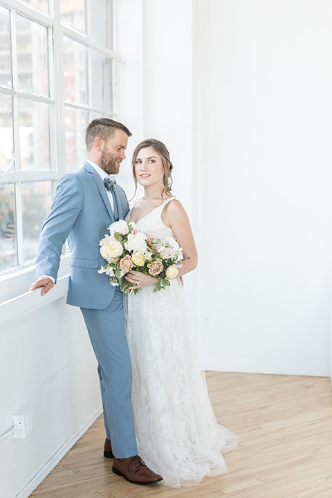 bride and groom by the windows, the bride in a flowing white gown with a lace detailing and straps.  The groom in a light blue suit with a grey velvet bow tie