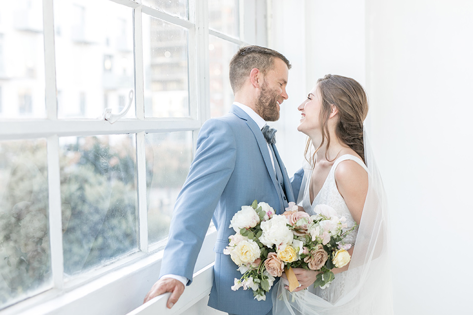  bride and groom by the windows looking at each other, the bride in a flowing white gown with a lace detailing and straps.  The groom in a light blue suit with a grey velvet bow tie 