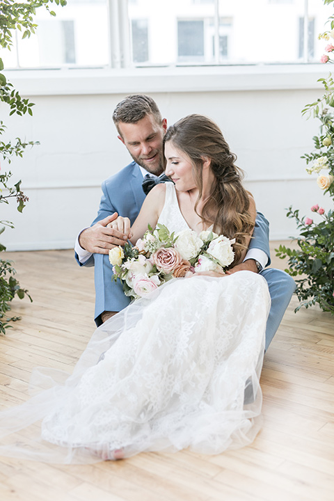  bride and groom sitting down holding each other, the bride in a flowing white gown with a lace detailing and straps.  The groom in a light blue suit with a grey velvet bow tie 