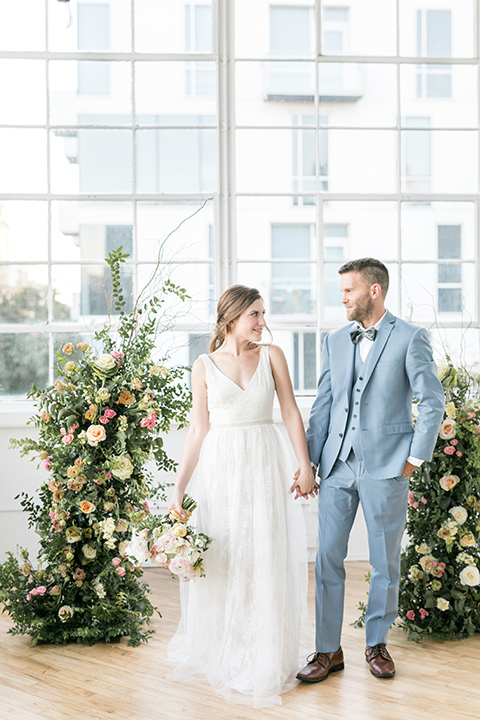  bride and groom at the ceremony, the bride in a flowing white gown with a lace detailing and straps.  The groom in a light blue suit with a grey velvet bow tie 