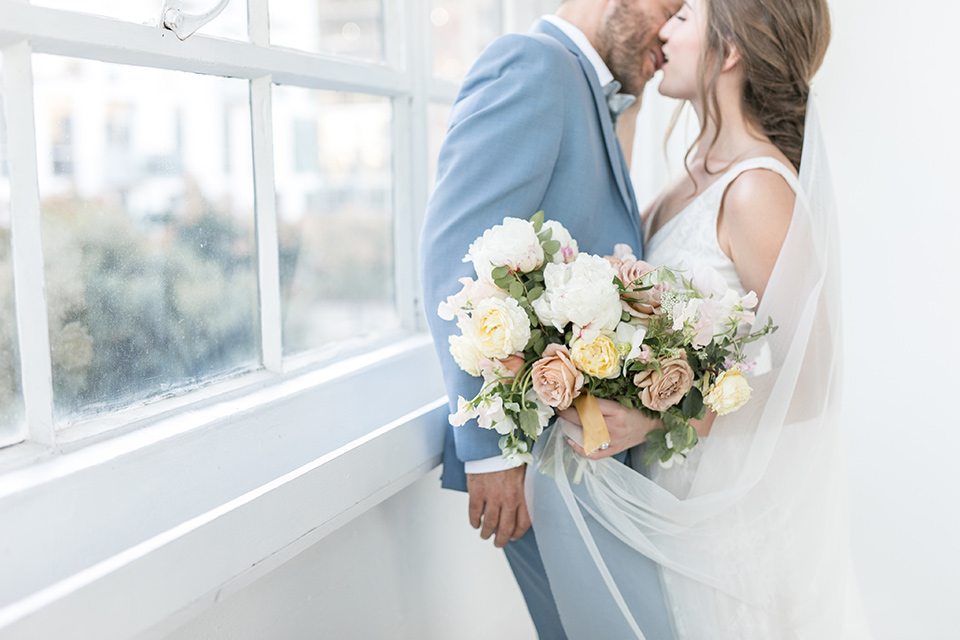  bride and groom by the windows looking at each other up close, the bride in a flowing white gown with a lace detailing and straps.  The groom in a light blue suit with a grey velvet bow tie 