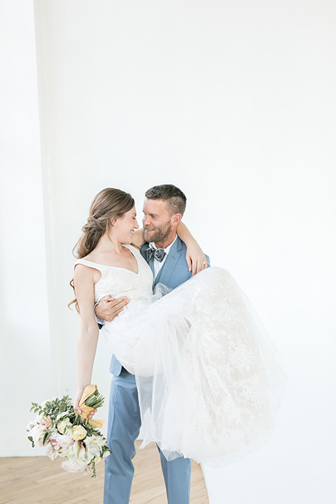  groom holding the bride in his arms, the bride in a flowing white gown with a lace detailing and straps.  The groom in a light blue suit with a grey velvet bow tie 