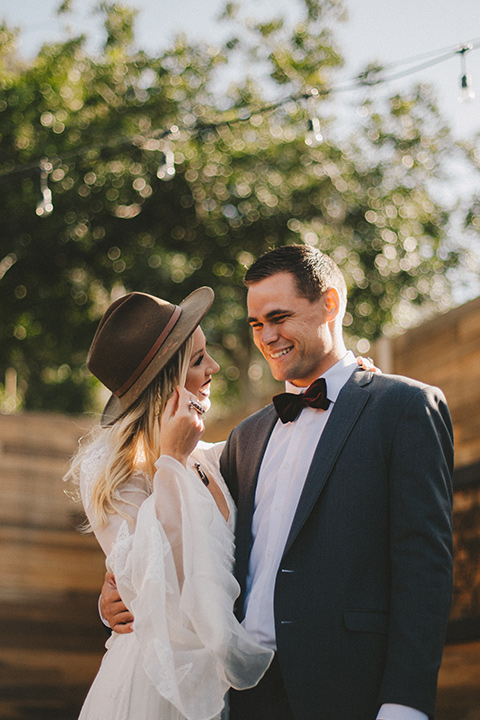  bride in a lace bohemian style gown with long sleeves and a wide brimmed hat and the groom in a dark grey notch lapel suit and a black bow tie
