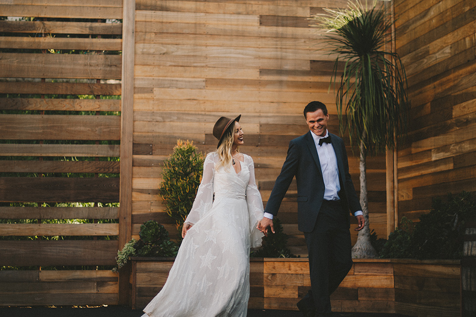  white lace gown with long billowing sleeves and a wide brimmed hat and the groom in a dark grey notch lapel suit and a black bow tie walking through venue