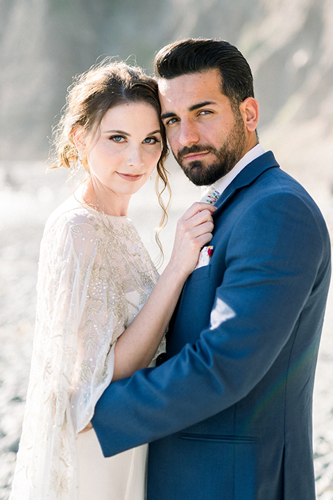  bride-in-a-flowing-gown-with-a-crystal-bodice-and-hair-in-a-loose-bun and the groom in a blue suit jacket with white pants and a floral long tie 