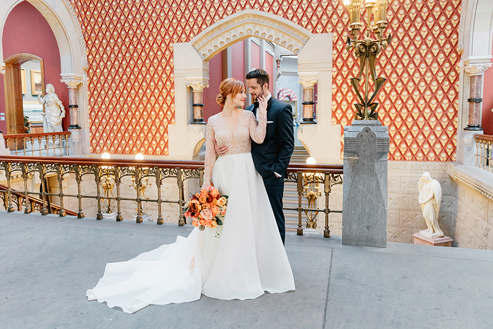  bride in a white ballgown with a crystal bodice and long sleeves with her hair in a low bun and the groom in a navy shawl lapel tuxedo with a black satin lapel standing in the museum