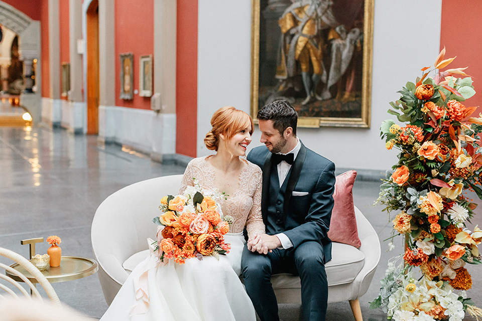  bride in a white ballgown with a crystal bodice and long sleeves with her hair in a low bun and the groom in a navy shawl lapel tuxedo with a black satin lapel sitting on a white couch