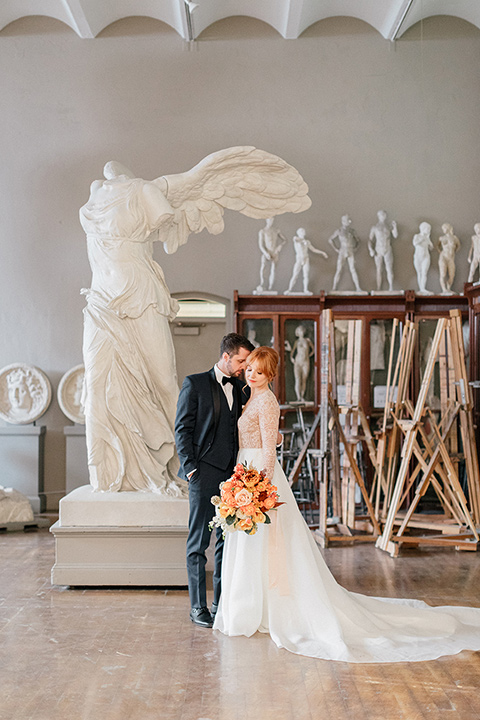  bride in a white ballgown with a crystal bodice and long sleeves with her hair in a low bun and the groom in a navy shawl lapel tuxedo with a black satin lapel near ancient sculptures