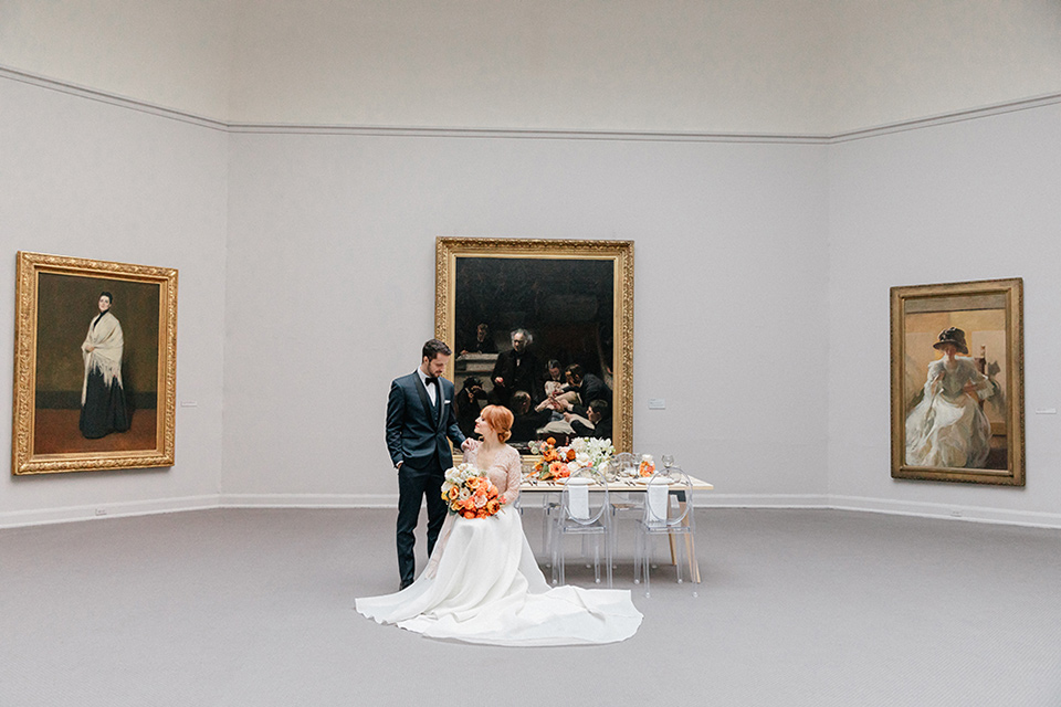  bride in a white ballgown with a crystal bodice and long sleeves with her hair in a low bun and the groom in a navy shawl lapel tuxedo with a black satin lapel sitting in the reception hall space