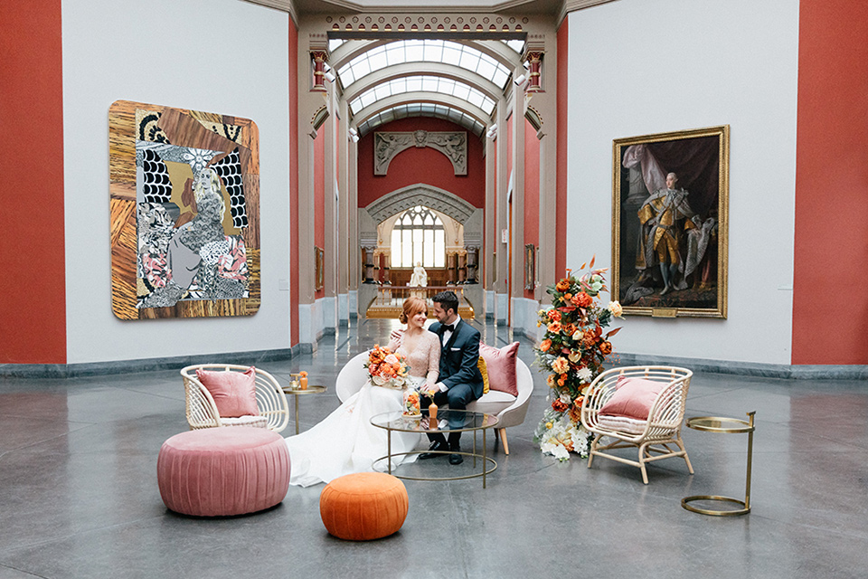  bride in a white ballgown with a crystal bodice and long sleeves with her hair in a low bun and the groom in a navy shawl lapel tuxedo with a black satin lapel sitting in the reception furniture space with white couches and pink velvet poofs