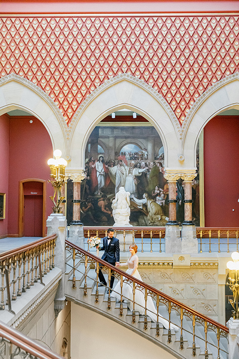  bride in a white ballgown with a crystal bodice and long sleeves with her hair in a low bun and the groom in a navy blue tuxedo with black shawl lapel design and black bow tie walking up the stairs