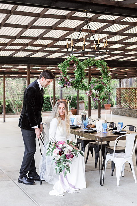 bride in a white lace gown and long sleeves and the groom in a black velvet tuxedo and a black long tie sitting together at the sweetheart table